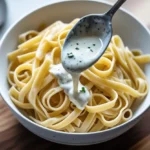 A bowl of fettuccine pasta topped with creamy white sauce being drizzled from a spoon